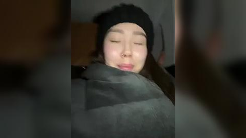 Media: A video of a young Asian woman with light skin, closed eyes, and a black beanie, wrapped in a gray blanket. The background is dimly lit, with blurred, indistinct objects.