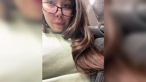 Media: Video of a young woman with long brown hair, wearing glasses and a ribbed green sweater, smiling slightly, taken from a low angle.