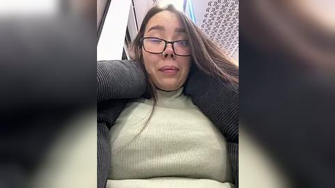 Media: Video of a young woman with glasses, fair skin, and long brown hair, wearing a gray sweater over a light green turtleneck, seated in a car with a blurry, dark background.