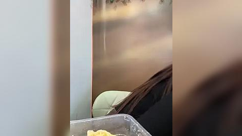 Media: A blurry video of a person\u2019s legs and a clear plastic container filled with food on a light-colored table, partially obscured by a wall. The background shows a blurred, possibly outdoor scene with trees and foliage.