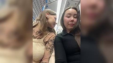 Media: Video of two women, one with long hair and the other with short hair, sitting on a bus. The woman with long hair wears a beige sweater with intricate designs. The woman with short hair is dressed in a black top.