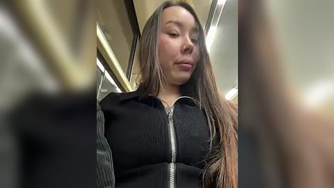 Media: Video of a young Asian woman with long brown hair, wearing a black ribbed sweater, standing on a subway train with a blurred background and fluorescent lighting.