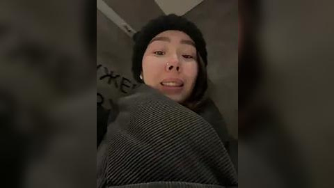 Media: A video of a young woman with light skin, long dark hair, and a black beanie, smiling, wearing a gray sweater, taken from a low angle, indoors, dim lighting.