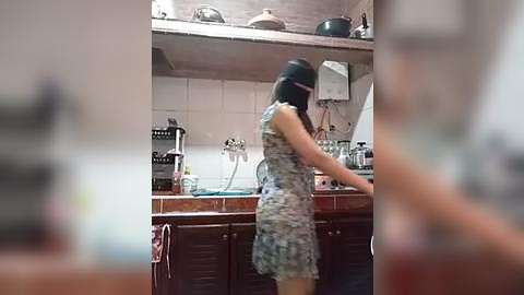 Media: Video of a woman with medium-length dark hair wearing a black headband and a floral-patterned dress, washing dishes in a rustic kitchen with wooden cabinets, tiled backsplash, and various kitchen utensils.