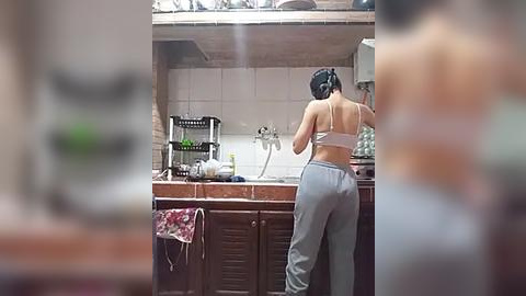 Media: Video of a woman in a kitchen with a wooden countertop, wearing a white bra and gray sweatpants, washing dishes. Background includes a sink, metal rack, and beige tiles.