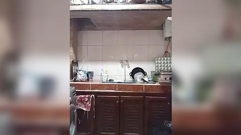 Media: Video of a small, cluttered kitchen with beige-tiled walls, a wooden sink, a cluttered countertop, and a black kettle.