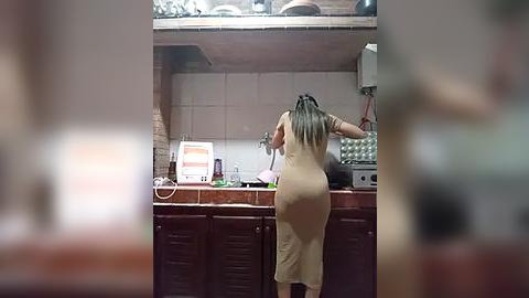 Media: A video shows a nude woman with long black hair, facing away, washing dishes in a small, dimly-lit kitchen with tiled walls and a countertop cluttered with dishes and appliances.