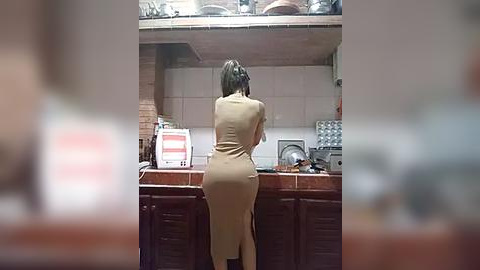 Media: Video of a woman with medium-length brown hair, wearing a tight, beige dress, standing in a small, cluttered kitchen with tiled walls and wooden cabinets.