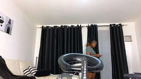 Media: Video of a modern, minimalist bedroom with white walls, black curtains, a black leather chair, and a woman with dreadlocks wearing a grey top and shorts.