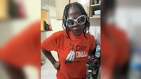 Media: Video of a middle-aged Black woman with short dreadlocks, wearing oversized white sunglasses and an orange t-shirt with a black eagle design. She stands in a cluttered kitchen with cabinets and countertops, holding a bottle.