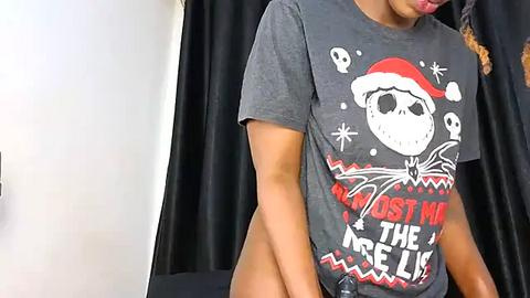 Media: Video of a woman with medium brown skin, wearing a gray holiday-themed t-shirt with a skull, wearing nothing underneath, standing against black curtains.