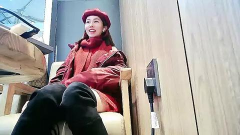 Media: Video of an East Asian woman with long brown hair, wearing a red beret, red turtleneck sweater, black thigh-high boots, and sitting in a modern, light-wooden chair inside a stylish, minimalist room with a glass door.