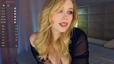 Media: Video of a blonde, fair-skinned woman with large breasts wearing a black sheer top, in a bedroom with a bed and a headboard.