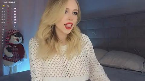 Media: Video of a blonde woman with wavy hair, wearing a white knitted sweater, smiling playfully, in a dimly lit bedroom with gray walls and a plush bed.