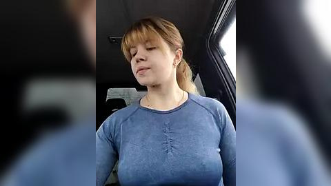 Media: Video of a young woman with light skin and brown hair, wearing a blue long-sleeve shirt, seated in a car. Her eyes are closed, and she appears relaxed.