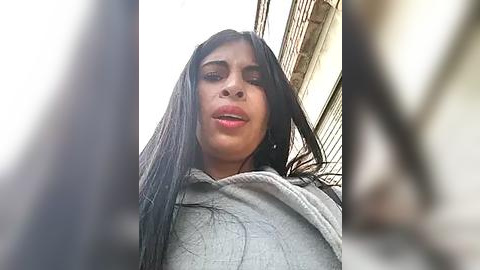 Media: Video of a young woman with long black hair, wearing a light gray sweater, taken from a low angle, partially blurred, in an outdoor setting with a building and power lines in the background.