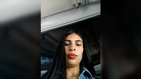 Media: Video of a young woman with long black hair, light brown skin, and full lips, wearing a denim jacket, taken from inside a car.