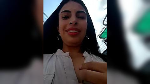 Media: A video of a smiling woman with medium skin tone, dark hair, and a white blouse, taken from a low angle, with blurred objects in the foreground.