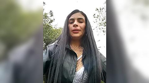 Media: Video of a woman with long, straight black hair, wearing a black leather jacket and a metallic sequin top, standing outdoors against a blurred greenery background.