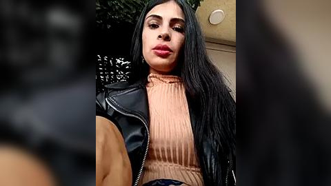 Media: A video of a Latina woman with long black hair, wearing a black leather jacket over a beige ribbed turtleneck, standing outdoors near a beige building.