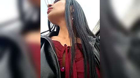 Media: A close-up video of a woman with long, straight black hair, wearing a black leather jacket over a maroon blouse, standing outdoors against a blurred background.