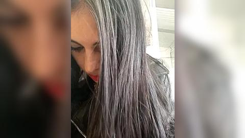 Media: Video of a woman with long, straight, silver hair in a black leather jacket, looking down. The background is blurred, focusing attention on her hair and attire.