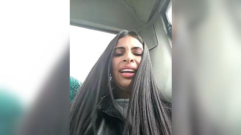 Media: Video of a woman with long, straight black hair, wearing a black leather jacket, smiling inside a car, surrounded by blurred, indistinct objects.