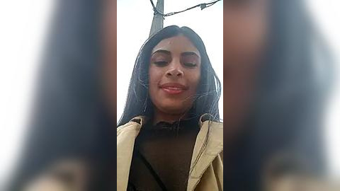 Media: Video of a young South Asian woman with long, straight black hair and medium-brown skin, wearing a beige jacket over a brown shirt. The background is blurred, featuring an overcast sky.