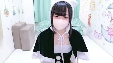 Media: Video of a young Asian woman in a black and white maid costume with a white mask, standing in a pastel-colored bedroom with floral wallpaper and a green bedspread.