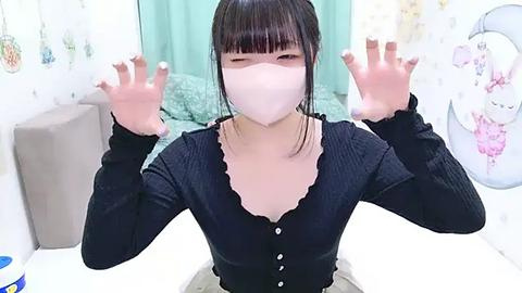 Media: Video of an Asian woman with straight black hair, wearing a black ribbed cardigan, white mask, and face shield, making a peace sign in a white room with teal curtains and floral wallpaper.