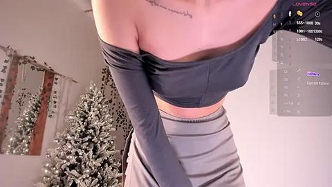 Media: Video of a woman with light skin and a small tattoo on her collarbone, wearing a black off-the-shoulder crop top and gray high-waisted pants, standing in a room with a Christmas tree and a mirror.