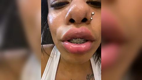 Media: Video of an Asian woman with full, glossy pink lips and metal braces, wearing a white top. Her skin is smooth, and she has a small nose piercing. The background is blurry, focusing on her face.