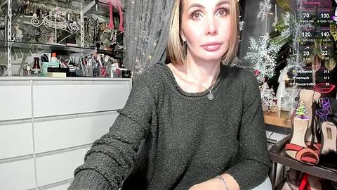 Media: Video of a woman with shoulder-length blonde hair, wearing a gray knit sweater, sitting in a cluttered, well-lit room filled with makeup brushes, clothing, and a digital clock displaying a time of 10:02.