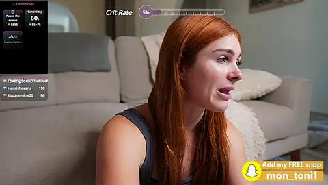 Media: Video of a red-haired woman with a fair complexion, wearing a sleeveless gray top, sitting on a couch in a living room with a beige sofa and a white pillow.