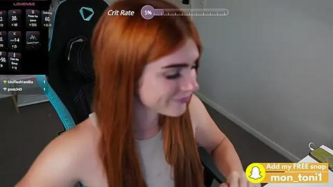 Media: Video of a young red-haired woman with pale skin, wearing a white tank top, seated in a black gaming chair with blue accents. Background includes a computer screen displaying a digital clock and various icons.