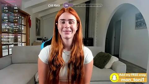 Media: Video of a fair-skinned woman with long, wavy red hair, wearing a white crop top, sitting in a modern living room with a grey couch and large windows.