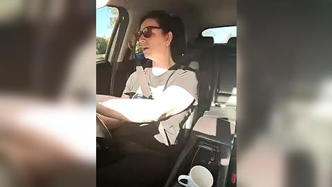 Media: Video of a woman with short dark hair and sunglasses, wearing a white blouse and black pants, sitting in a car, holding a book and a white coffee cup.