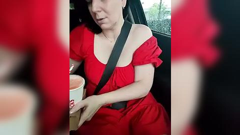 Media: A video of a woman in a red dress and black seatbelt, holding a pink cup, sitting in a car with blurry people in the foreground.