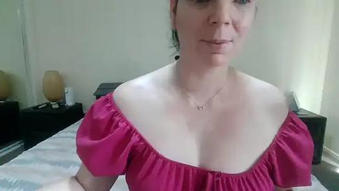 Media: Video of a fair-skinned woman with dark hair, wearing a low-cut pink top, standing in a bedroom with beige walls, a bed, and nightstands with lamps.