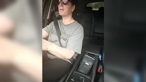 Media: Video of a middle-aged woman with short dark hair, wearing dark sunglasses and a grey t-shirt, driving a black car with a black interior. She has a shoulder strap on and is gripping the steering wheel.