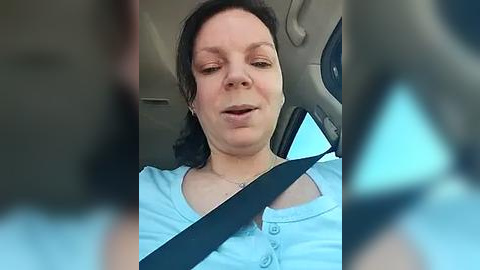 Media: A video of a woman with short brown hair, wearing a light blue shirt, driving with a seatbelt, blurred background, casual setting.