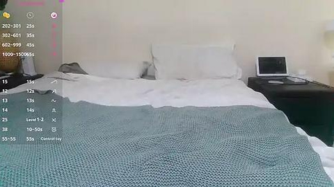 Media: A video of a neatly made bed with white linens and a teal knitted blanket. A white pillow rests against a light beige wall. A black nightstand with a closed laptop is on the right.