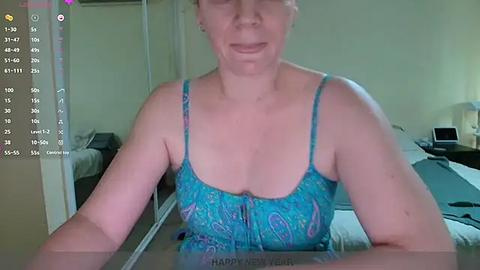 Media: A video of a smiling, middle-aged woman with fair skin and blonde hair, wearing a turquoise, patterned spaghetti-strap tank top. She is indoors, in a dimly lit bedroom with a bed, nightstand, and mirror in the background.