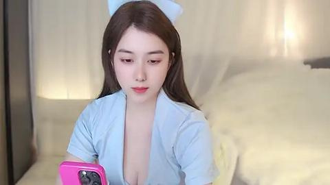 Media: Video of an East Asian woman with long brown hair, wearing a blue robe, standing in a softly lit bedroom, holding a pink phone.