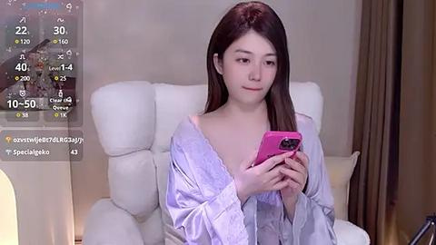 Media: Video of an Asian woman with long brown hair, fair skin, wearing a pink phone case, light robe, sitting on a white chair.