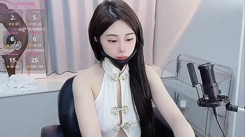 Media: Video of an East Asian woman with long black hair, wearing a white halter top with gold buttons, sitting in a chair with a black mask over her mouth. Background features a wall with a clock, a hairdryer, and a mirror.