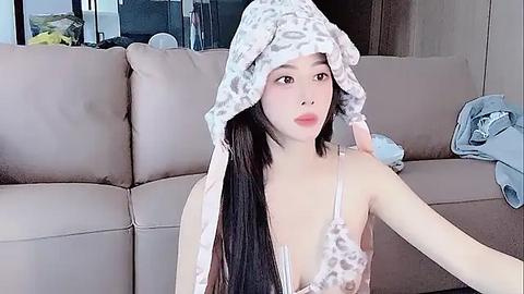 Media: Video of an Asian woman with long black hair, wearing a leopard-print headband, light pink lingerie, and sitting on a beige sofa. Background includes a partially visible window and a blue garment on the right.