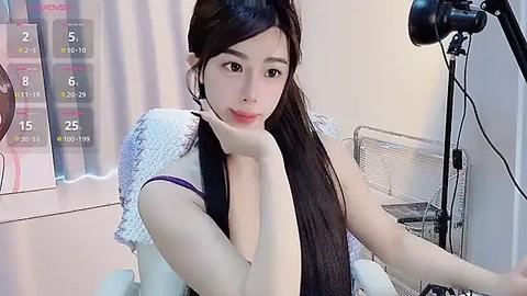 Media: Video of an East Asian woman with long black hair, light skin, and a slender build, sitting in a modern room, wearing a purple bralette, with a webcam on a stand in the background.