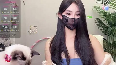 Media: Video of an Asian woman with long black hair wearing a black mask, blue top, and beige fur stole, standing next to a small white Pomeranian dog in a room with a green plant and a calendar with daily weather data.