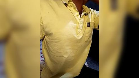 Media: A close-up video of a person wearing a yellow polo shirt with a badge on the left chest. The background is blurred, revealing a dark surface and a hint of a patterned fabric. The image is slightly out of focus.